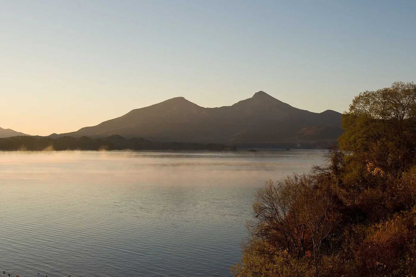 桧原湖の水面ごしに見る磐梯山。朝もやが湖面に立ち、手前の岸に紅葉した木が並ぶ