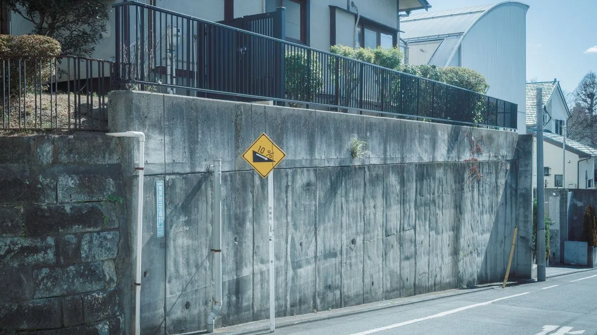 道路沿いのコンクリート擁壁の上に柵が立ち、その奥に住宅が建っている