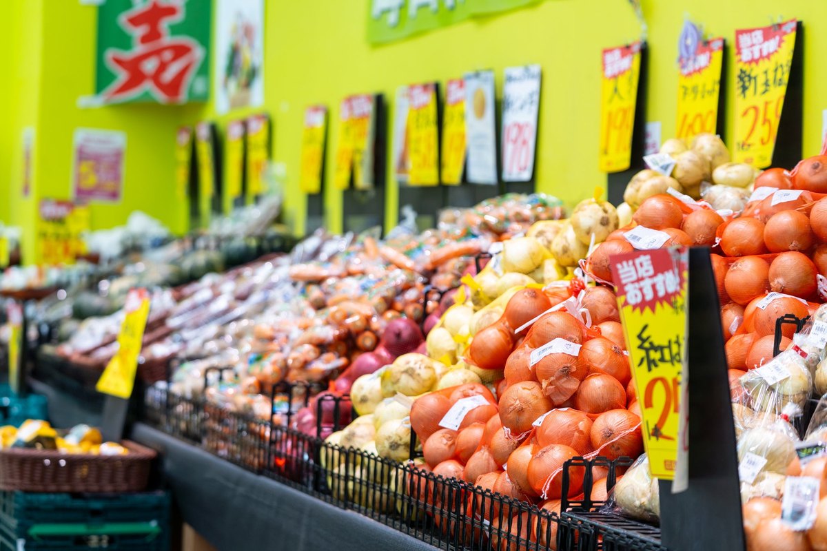 業務スーパーの青果売場。玉ねぎやじゃがいもが袋詰めで並んでいる