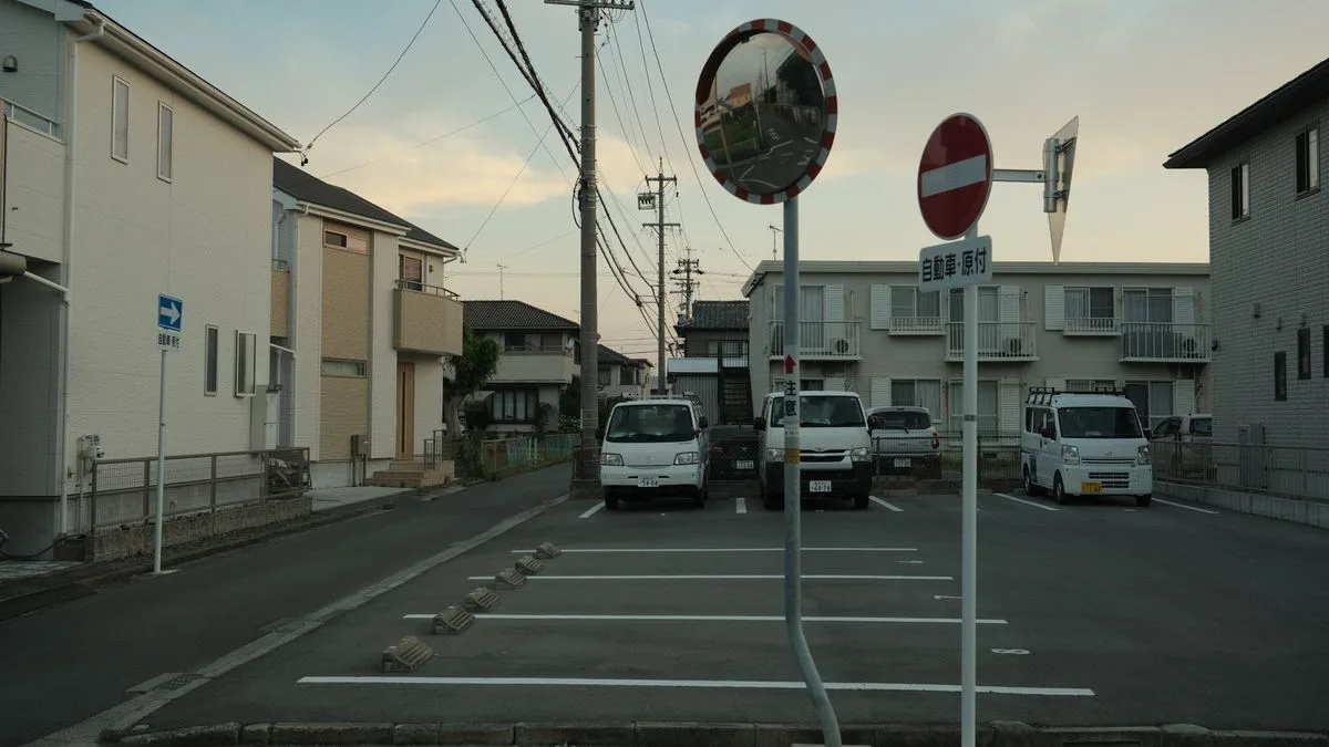 夕方の住宅街。電柱とカーブミラーの先に低層のアパートが並び、区画された駐車場が広がっている