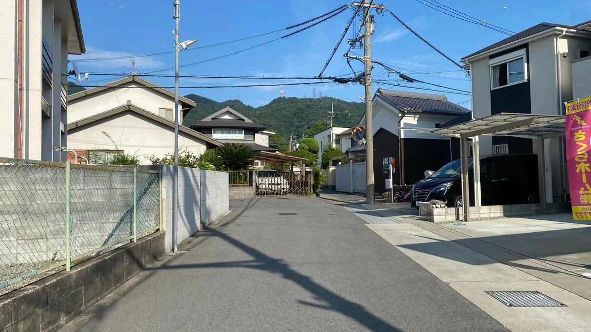 山を背にした静かな住宅街の道路。戸建てが並び、車が停まっている