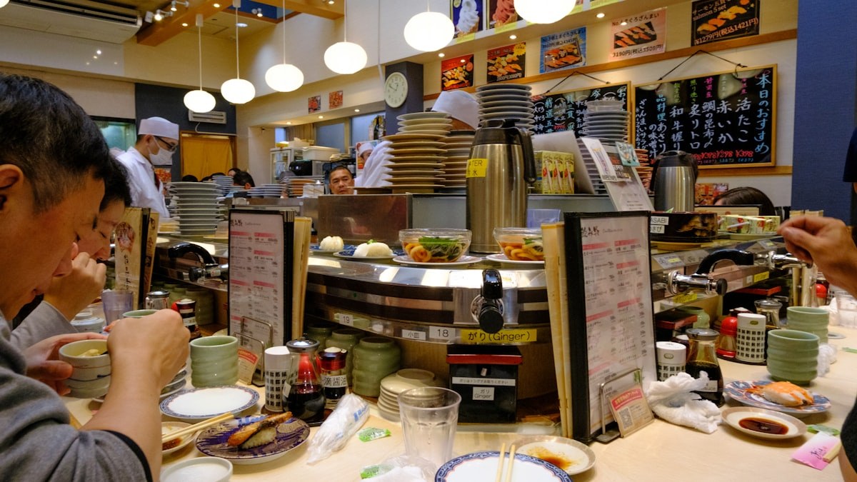 回転寿司店のカウンターで食事をする人たちと、壁に並んだ価格の書かれたメニュー