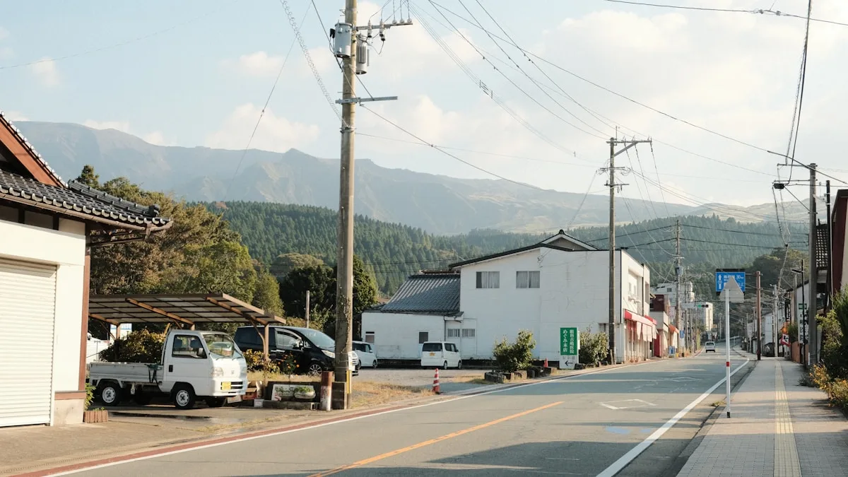 山を背にした地方の住宅地の通り。道沿いに住宅が建ち、軽トラックが停まっている