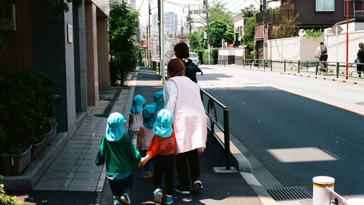 街の歩道を、帽子をかぶった小さな子どもたちが大人に付き添われて歩いている