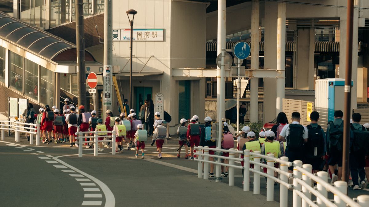 駅前の歩道を、帽子をかぶりランドセルを背負った小学生の列が歩いていく