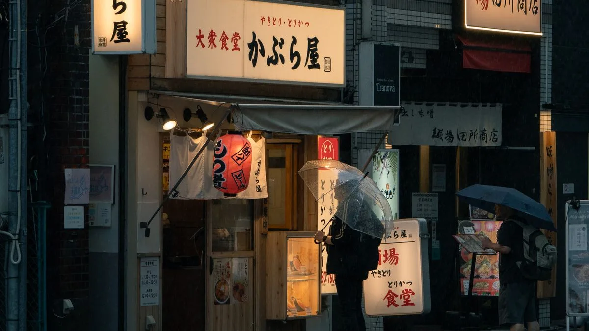 雨の降る日本の商店街。傘をさした人が店先に立っている