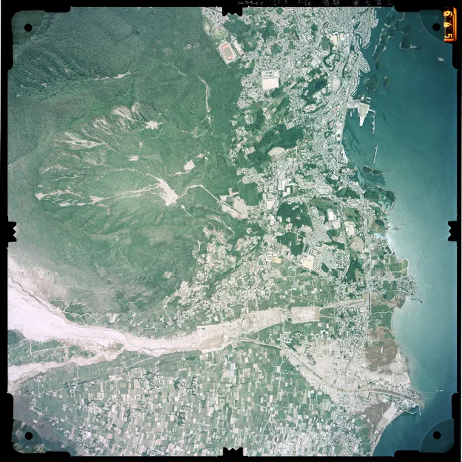 島原の上空から撮った空中写真。右上に有明海と市街地、左上に眉山の緑があり、画面の下側を山から海へ向かって白い帯が横切っている。これが火砕流の堆積物で埋まった水無川