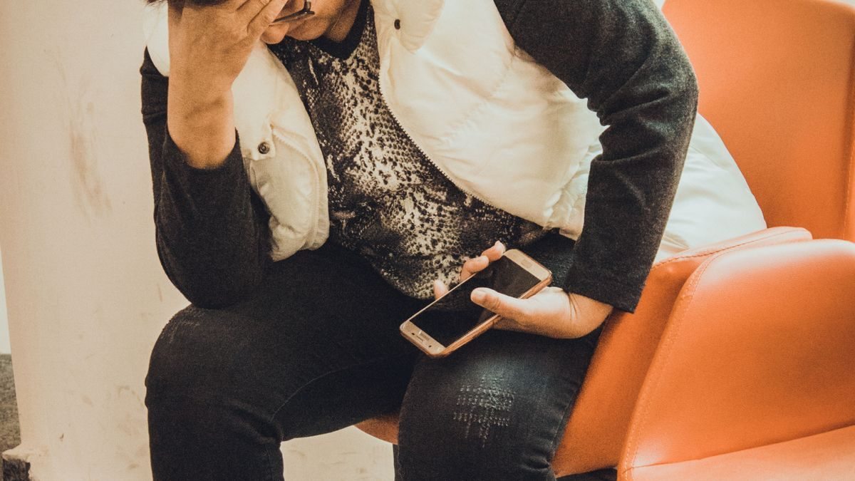オレンジ色のいすに座り、額に手を当ててうつむく人。もう一方の手にスマートフォンを握っている