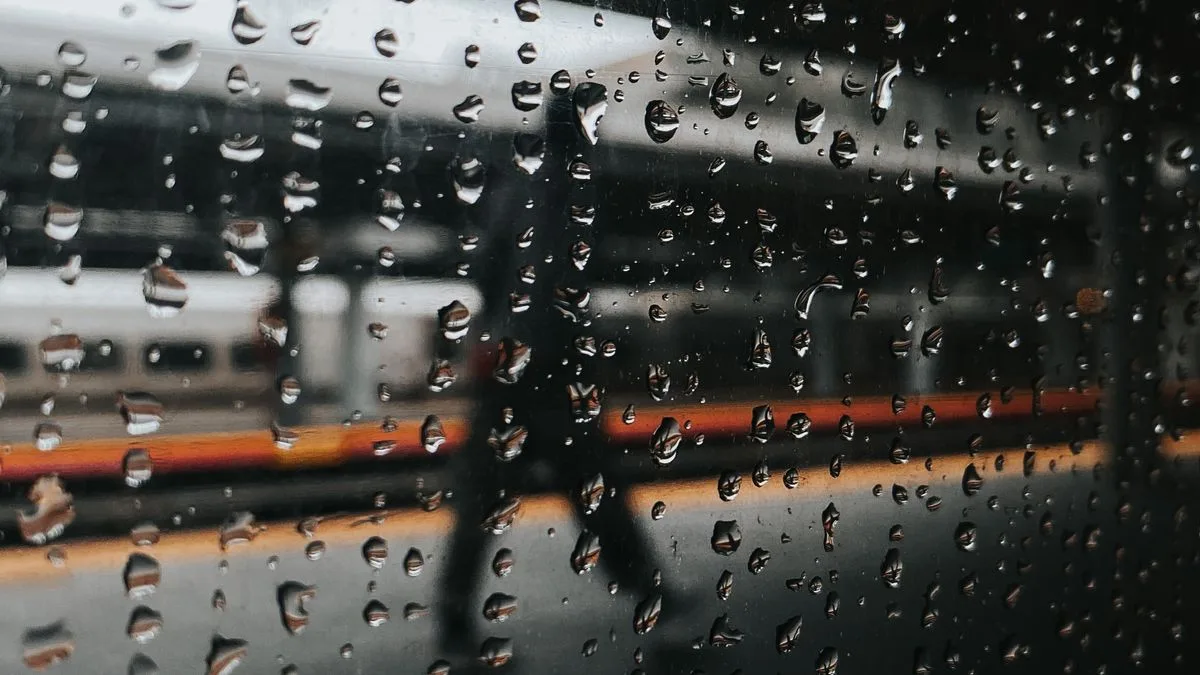 雨粒がついた窓越しに、停車中の列車のホームと歩く人のシルエットが見える