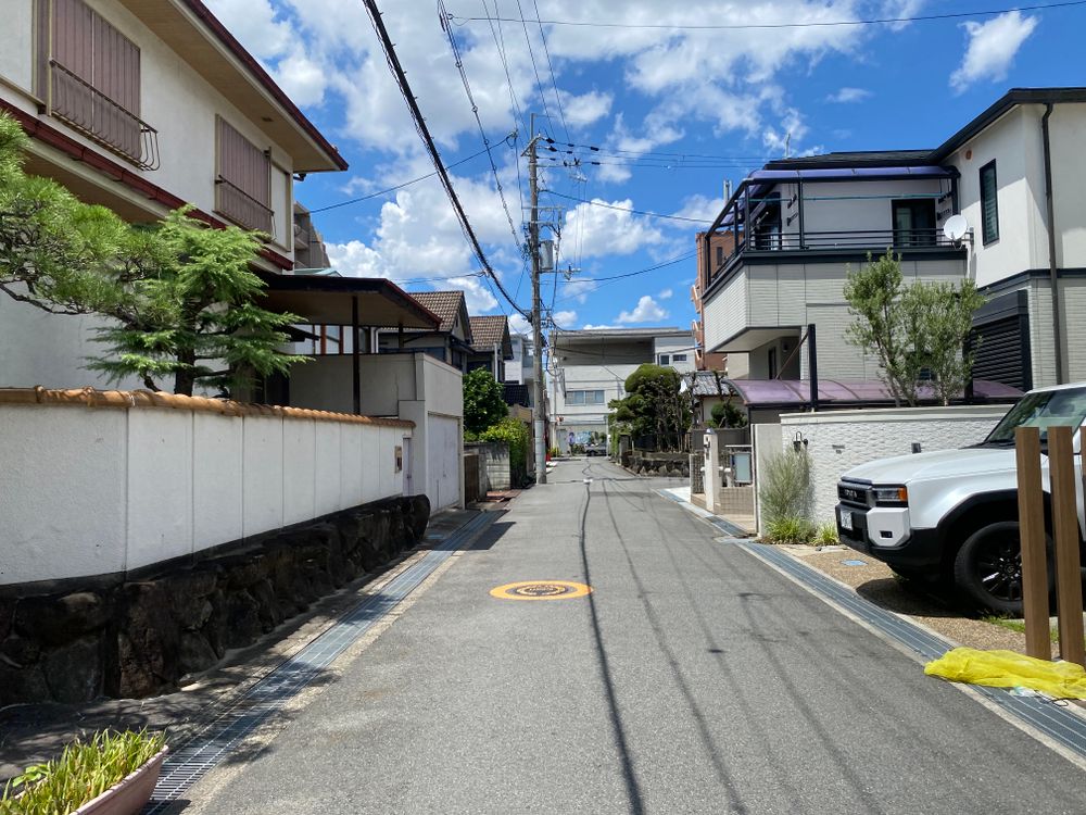 白い塀に沿った住宅地の道。新しめの戸建てが並び、奥に電柱と青空が見える