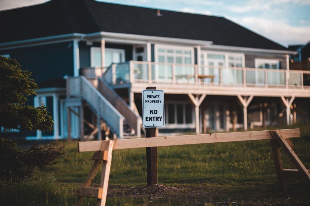 芝生の敷地の手前に木の柵と「PRIVATE PROPERTY NO ENTRY（私有地・立入禁止）」の看板が立ち、奥に住宅が見える