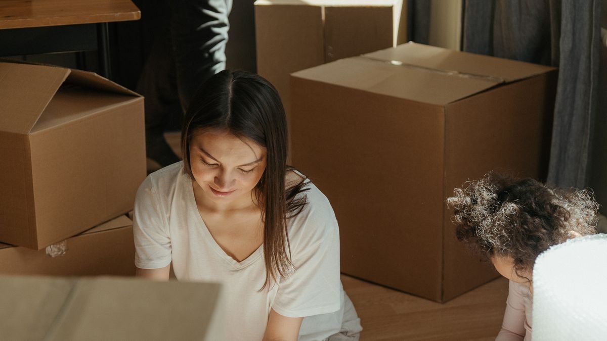 段ボール箱が積まれた部屋で、床に座って荷物を詰める女性と、その横にいる小さな子ども