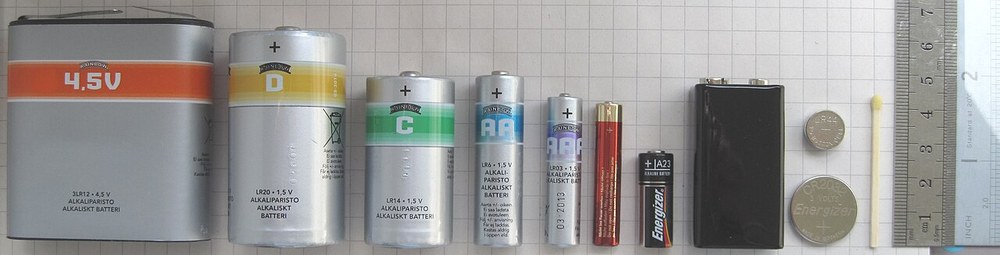 いろいろな電池を大きさの順に並べた写真。左から単1・単2・単3・単4などの乾電池、9ボルト電池、コイン形のCR2032、いちばん右の小さいのがアルカリボタン電池のLR44。マッチ棒と定規で大きさがわかる