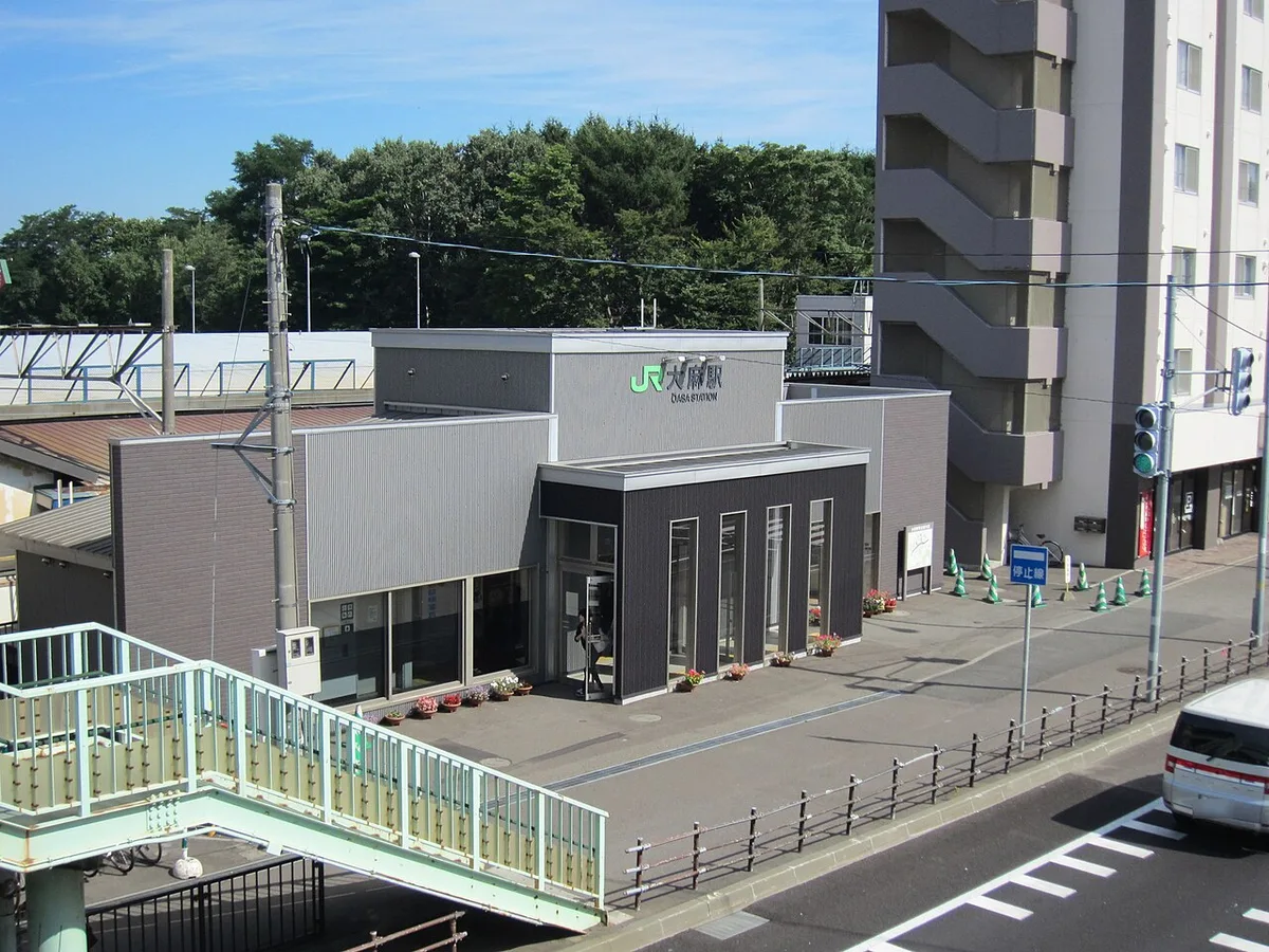 JR大麻駅の南口。グレーの駅舎の横にマンションが建ち、手前に歩道橋の階段がある