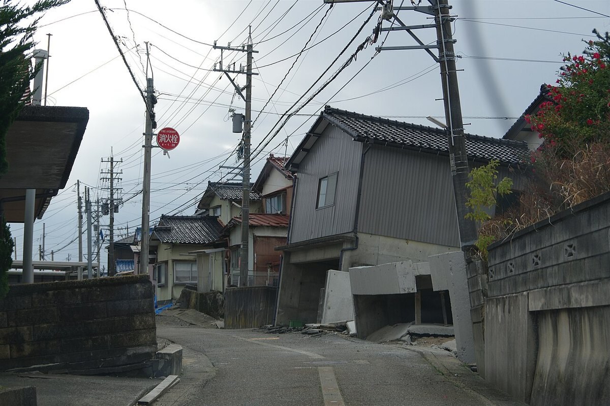 電柱が傾き、住宅の足元の車庫部分がずれて崩れている住宅街の坂道