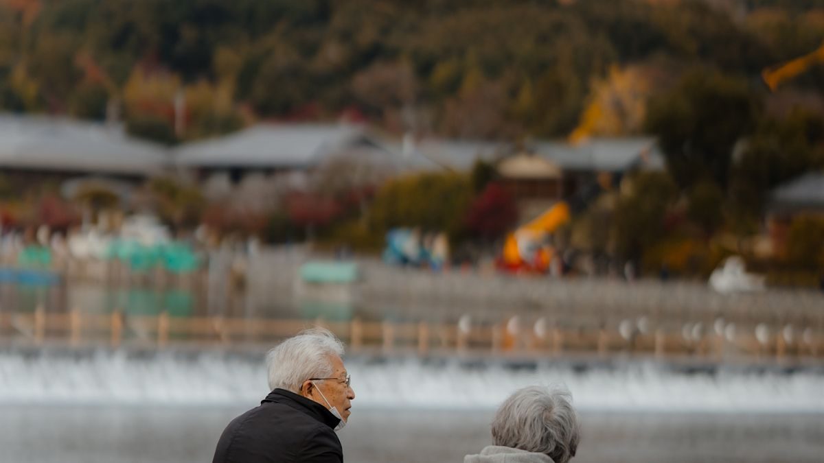 紅葉の山を背にした川辺で、白髪の男性と女性が並んで座り、川の流れを眺めている