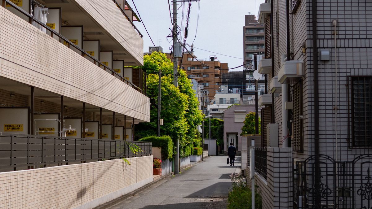 晴れた日の住宅街の細い道。片側にタイル張りの集合住宅、もう片側に戸建てが並び、奥に人が歩いている