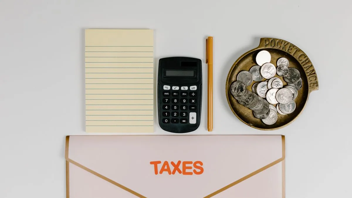 メモ帳と電卓とペン、硬貨をのせた皿、英語で「TAXES（税金）」と書かれた書類ばさみが並んだ机の上