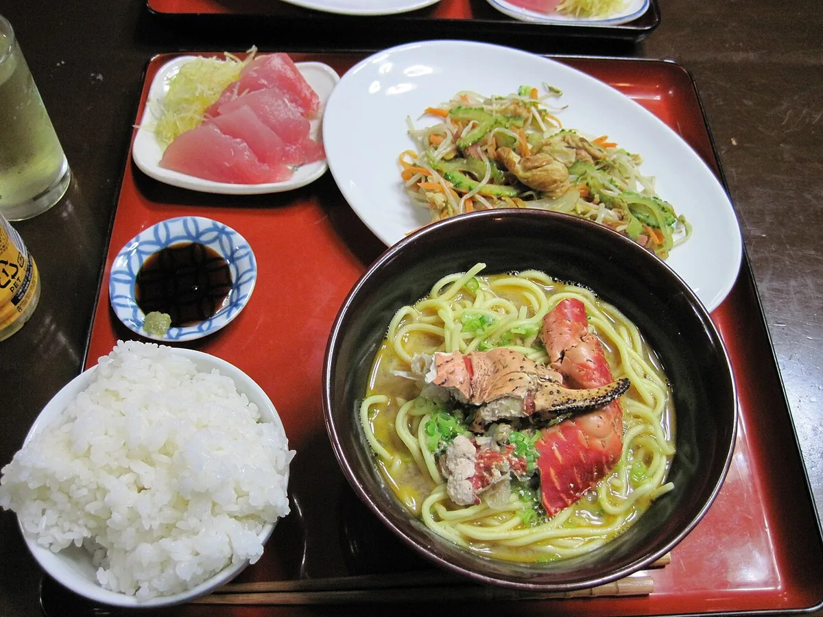 赤くゆで上がったヤシガニの脚とはさみがのった沖縄そば。横にご飯、刺身、ゴーヤーチャンプルー
