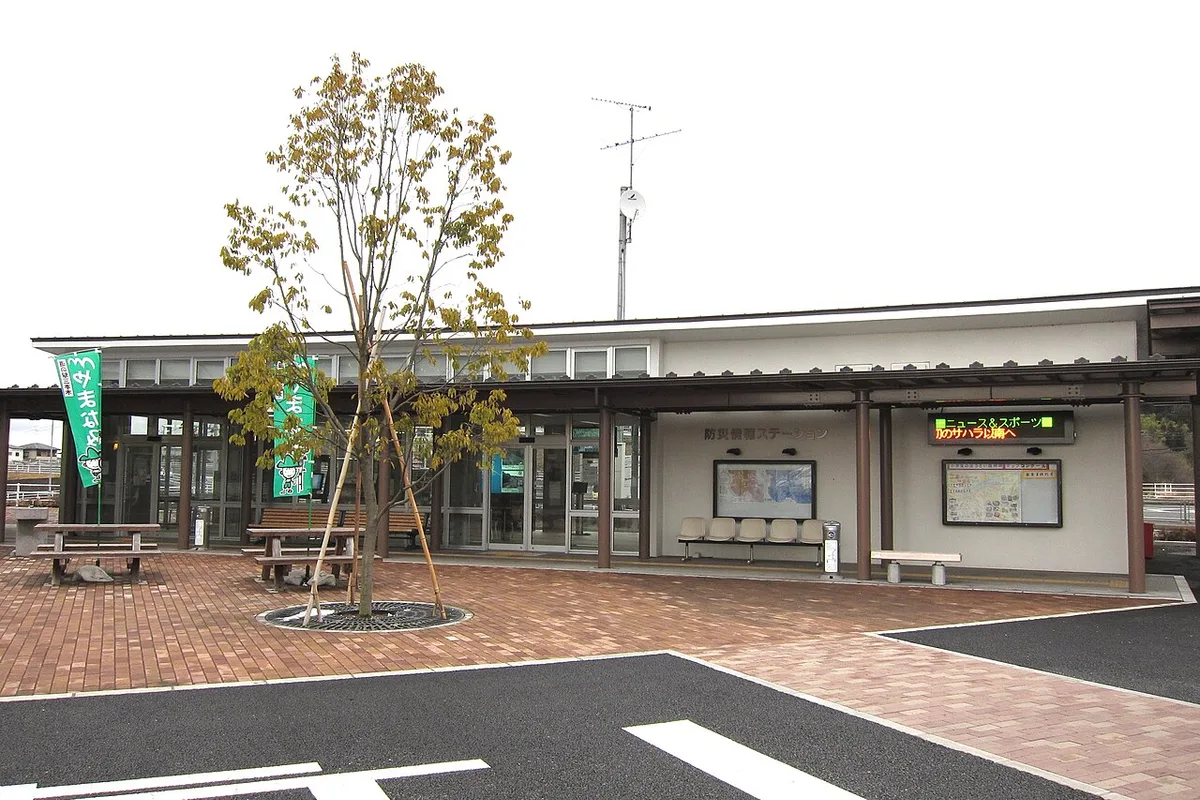 道の駅三本木の防災情報ステーション。平屋の建物の前にれんが敷きの広場と若い木がある