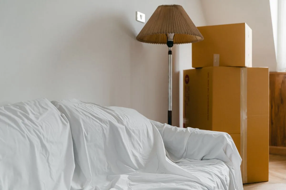 引っ越しの段ボールが積まれ、家具に白い布がかけられた部屋