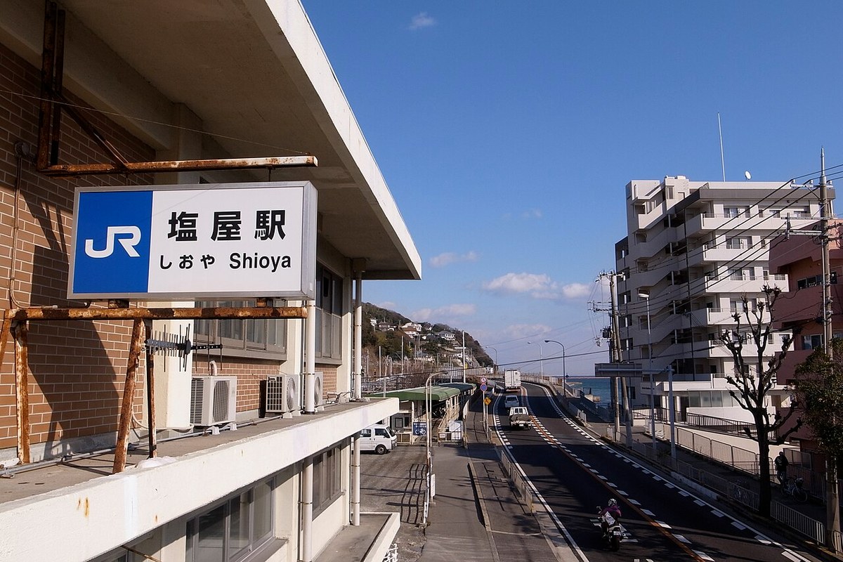 JR塩屋駅の駅名標と、海沿いの国道2号。奥の山の斜面に住宅が積み重なるように建っている