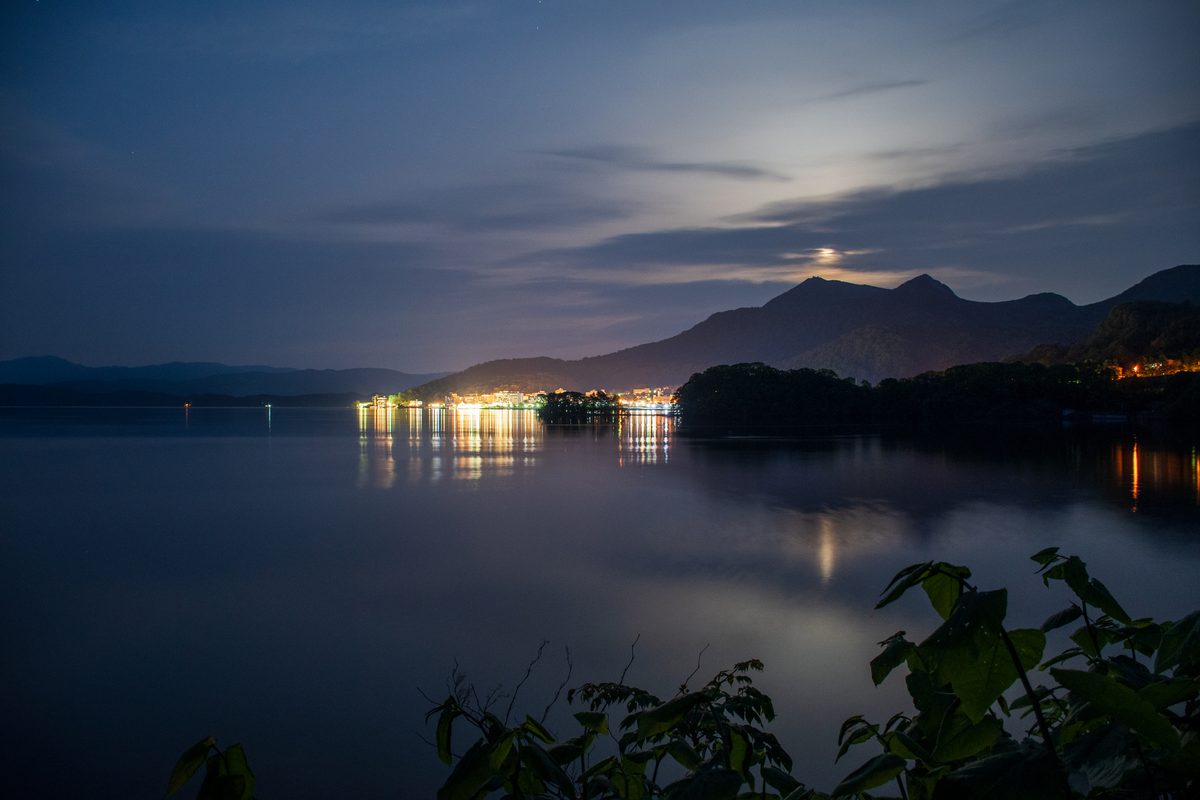 夜の洞爺湖。月明かりの下、湖岸に並ぶ街の明かりが湖面に映っている
