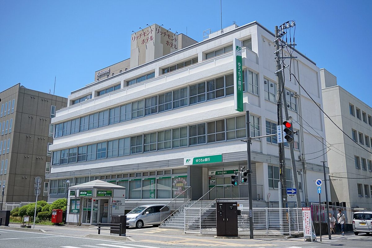 秋田市中通にあるゆうちょ銀行秋田店の建物。白い4階建てで、壁に緑の「ゆうちょ銀行」の看板があり、手前に銀行ATMのボックスと赤いポストがある